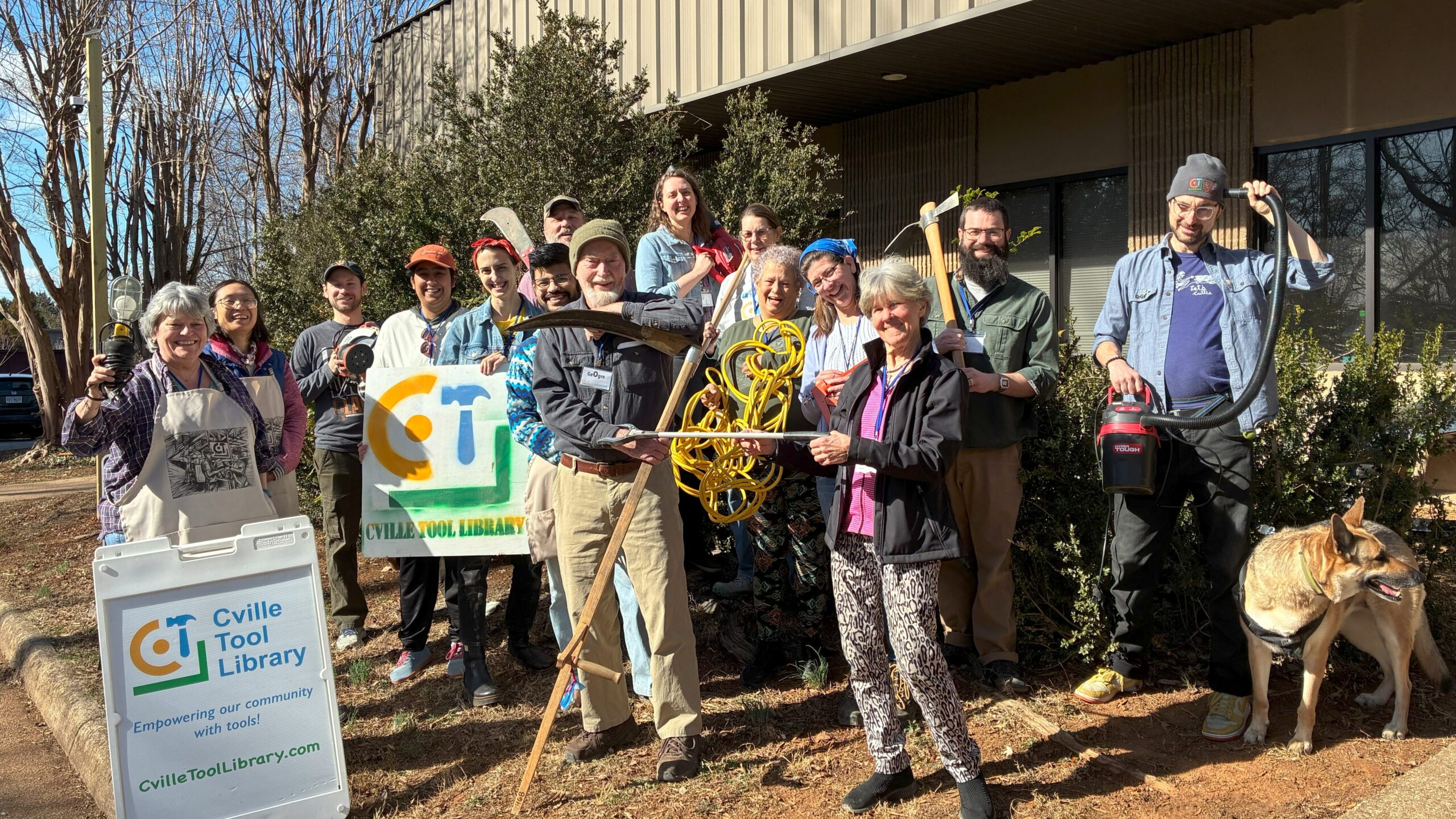 A large group of volunteers stands outside the entrance to the Cville Tool Library, holding various tools.